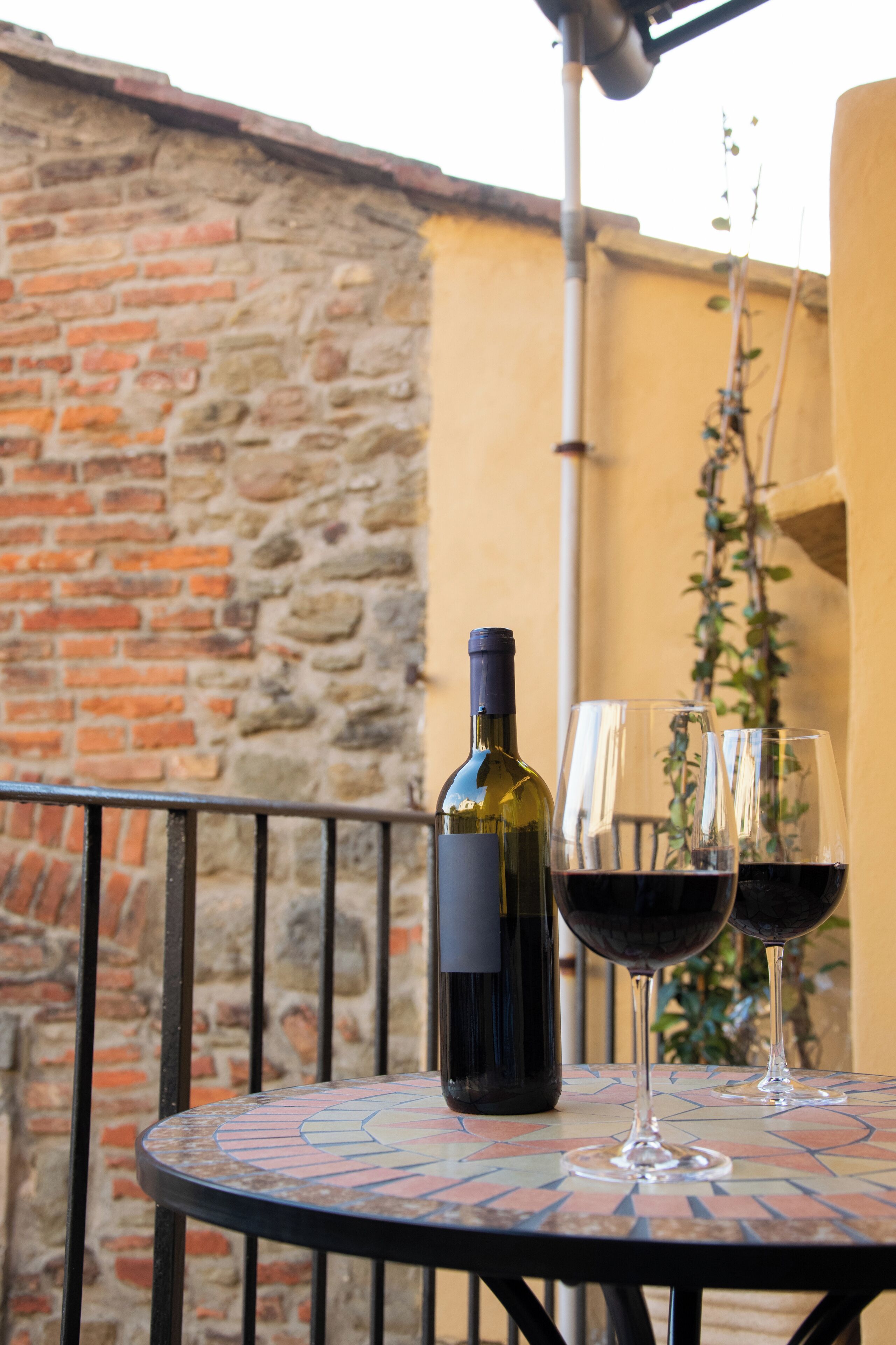 A table with a bottle of wine and two glasses on a tiled patio.