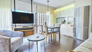 A modern living room with a flat-screen TV, a dining area with a table and chairs, a kitchen with a refrigerator, and a large window with a view of buildings.
