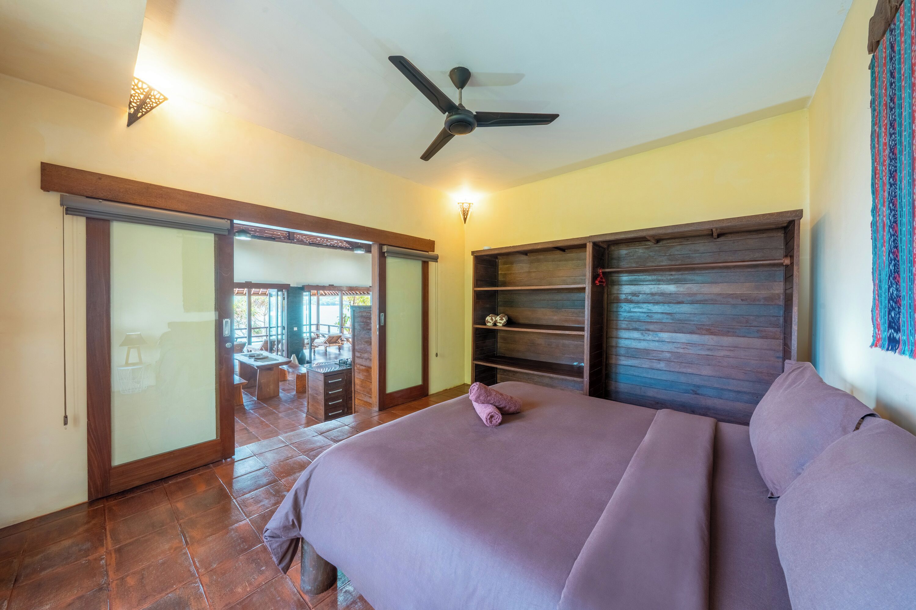 Un dormitorio con una cama, un ventilador de techo, una puerta corrediza de vidrio que da a un balcón y una estantería de madera.