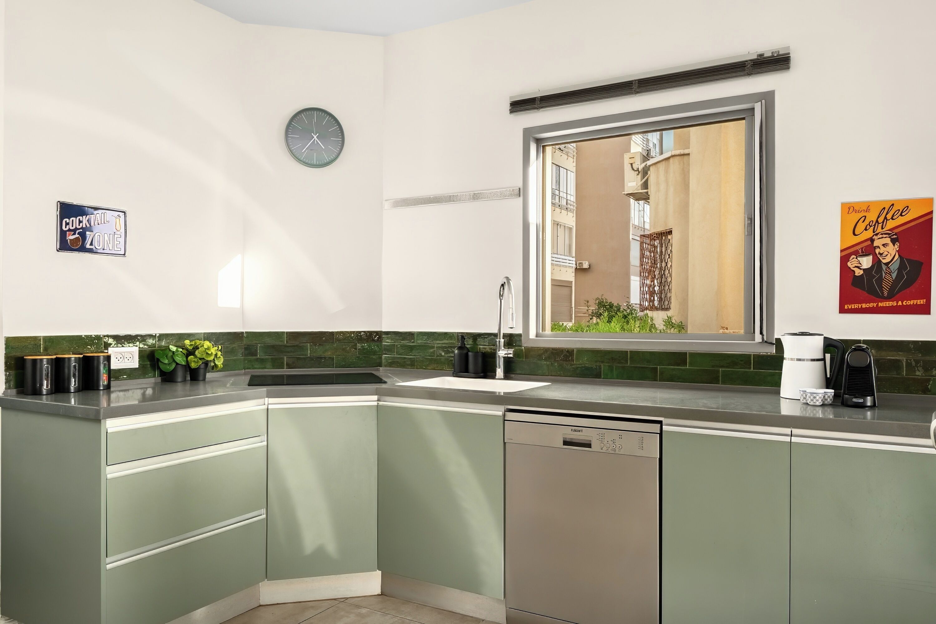 Een keuken met groene kastjes, een vaatwasser, een spoelbak, een klok en een raam met uitzicht op gebouwen.