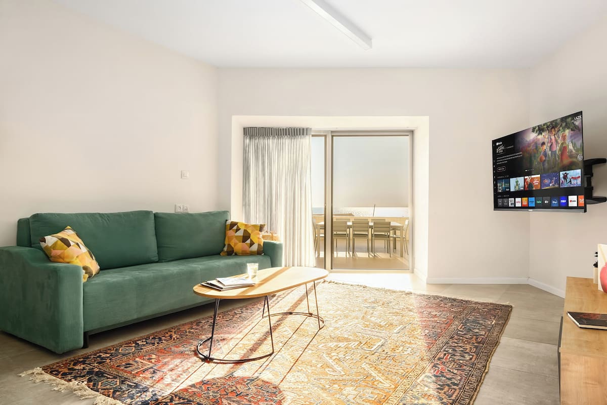A living room with a green sofa, a coffee table, a patterned rug, and a TV mounted on the wall.