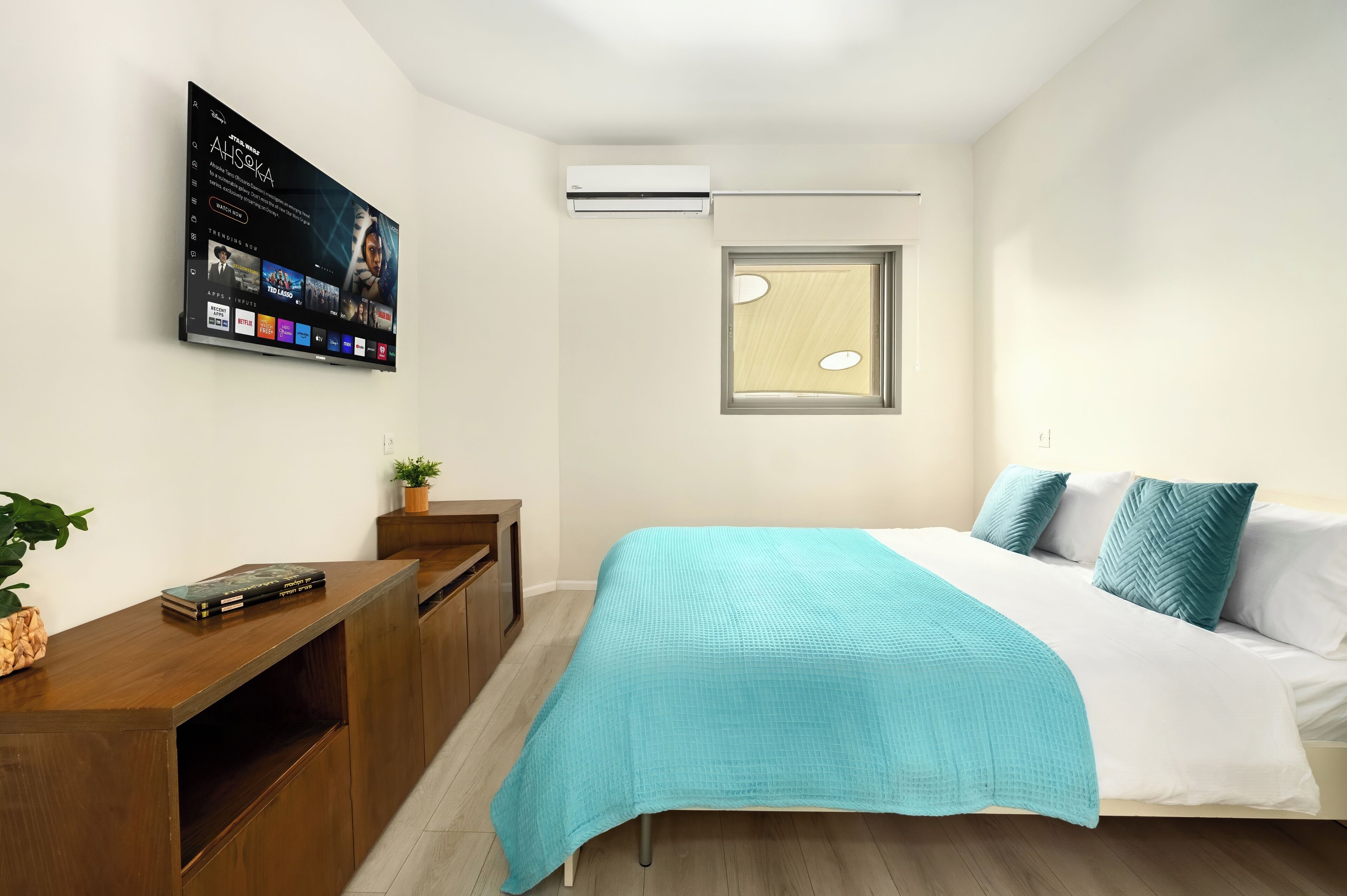 A bedroom with a bed, a TV mounted on the wall, a window, and a wooden desk with a plant.