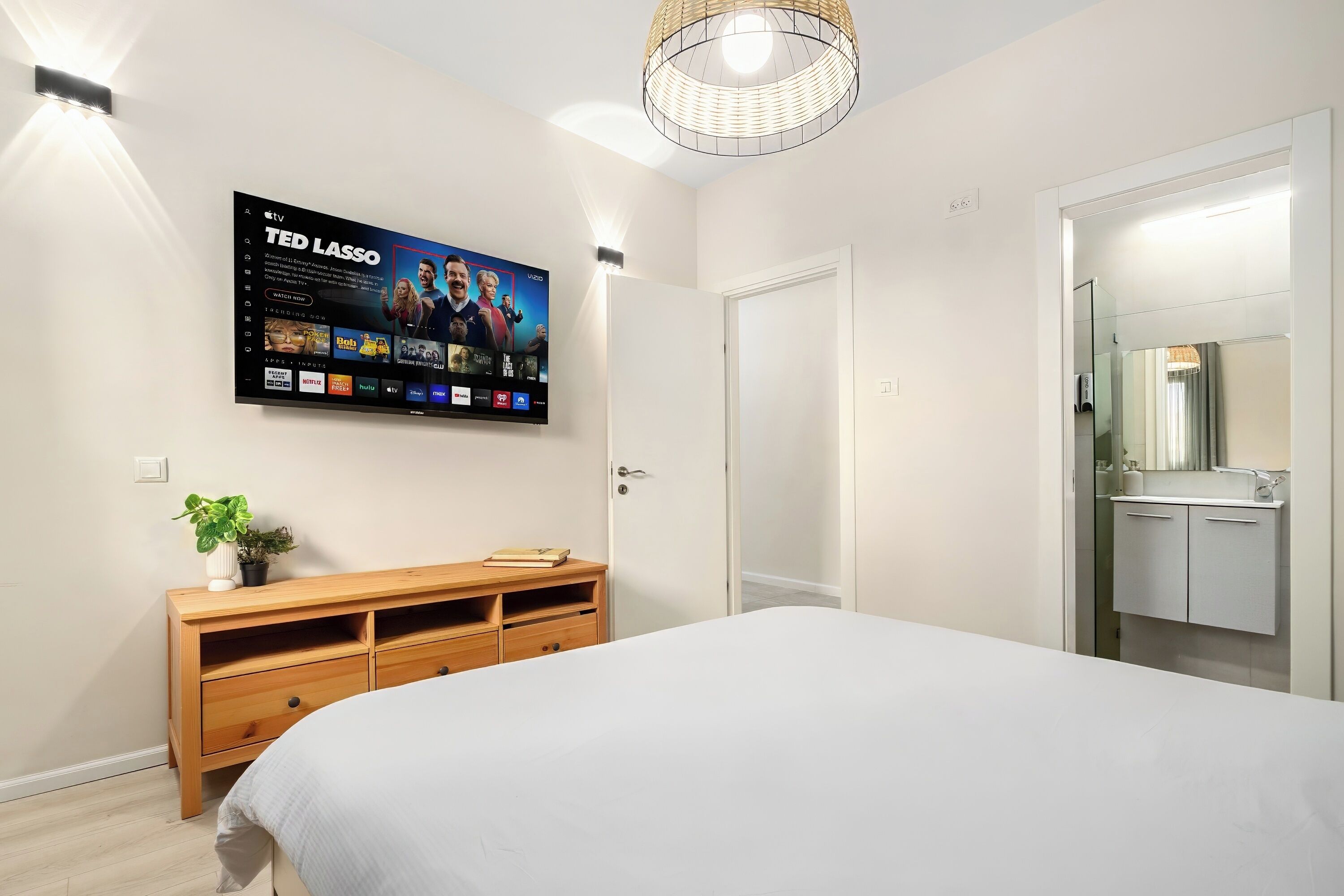 Un dormitorio con una cama, un televisor en la pared mostrando un programa, un aparador de madera y una puerta que da al baño.