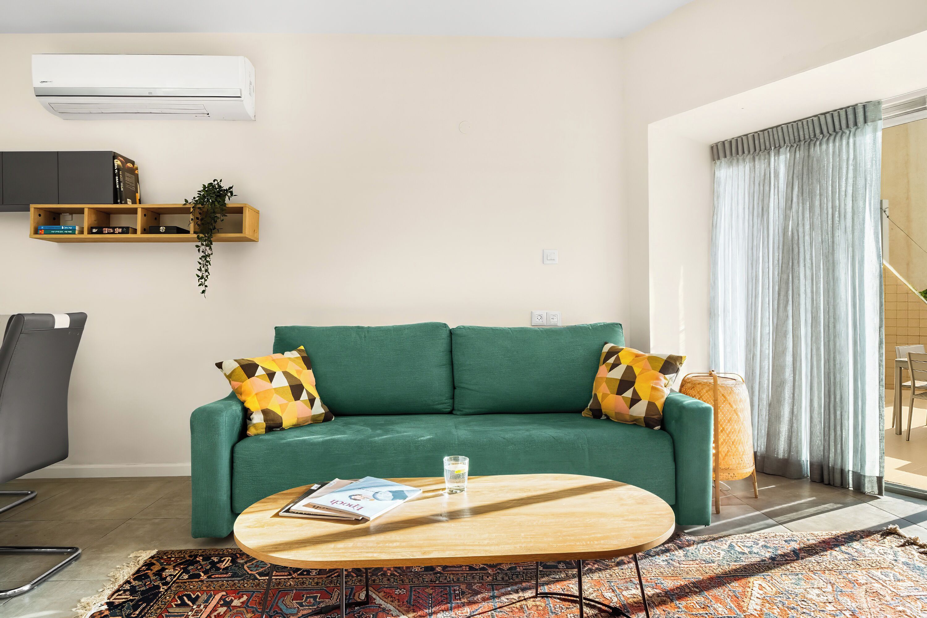 Un sofá verde con cojines estampados, una mesa de centro de madera con una revista, una mesita auxiliar de mimbre y un ventanal grande con cortinas.