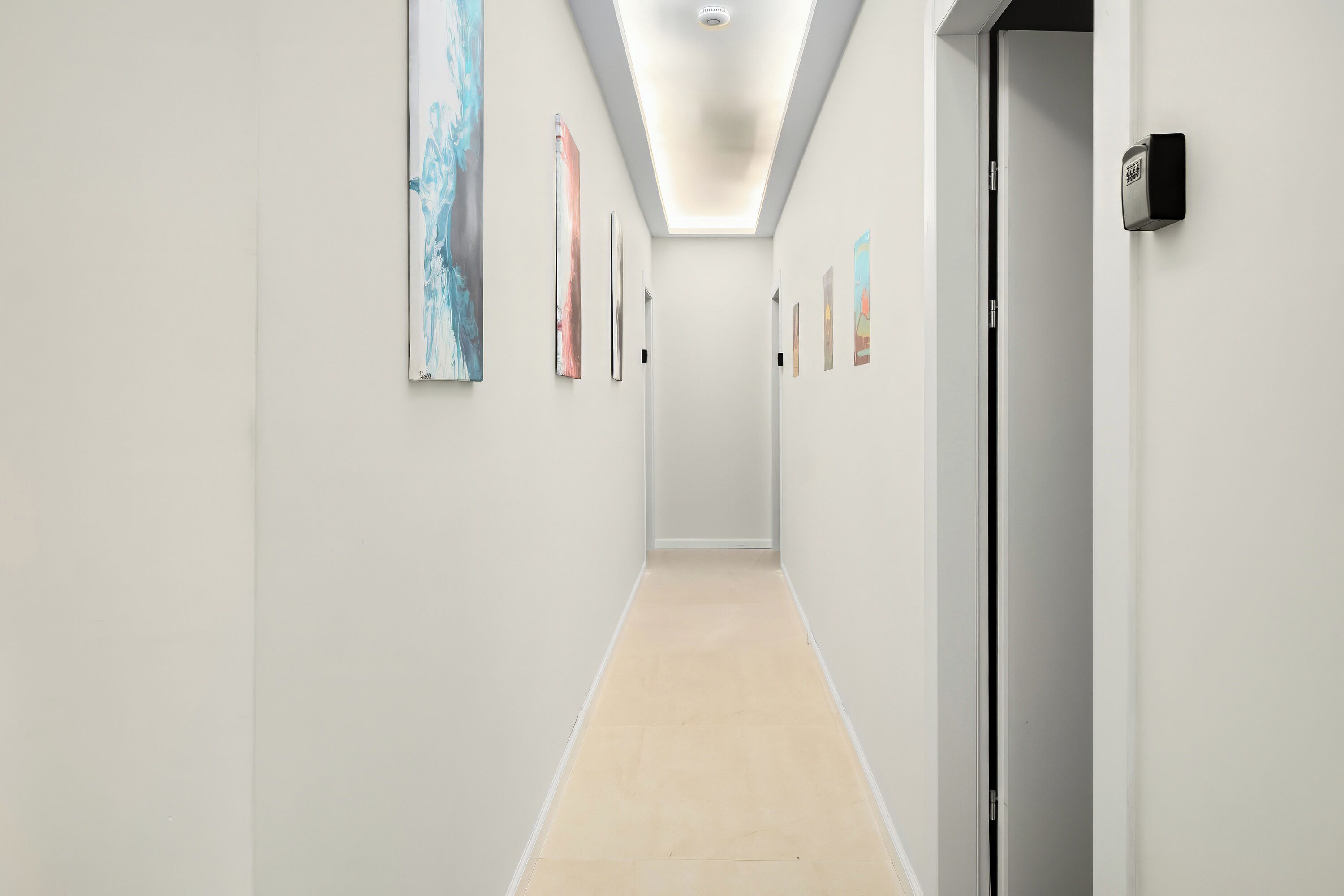 A modern hallway with light-colored walls, a tiled floor, and several framed artworks on the walls.