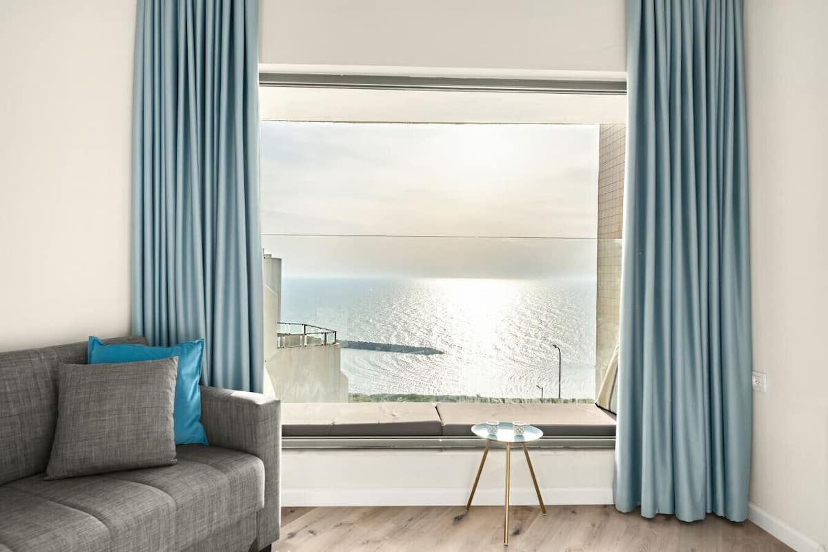 A modern living room with a grey sofa, a small round table, and a large window with a view of the sea.