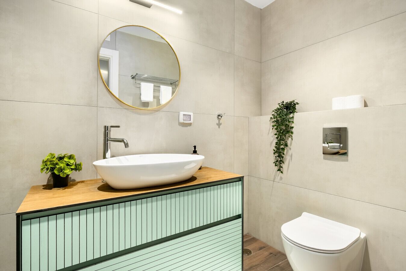 A modern bathroom with a large oval sink, a round mirror, a toilet, and a small potted plant.