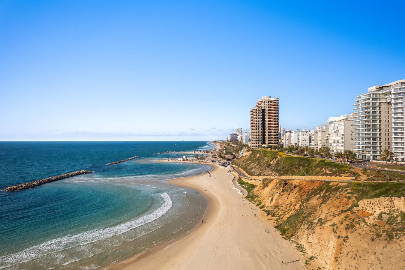 A coastal cityscape with a sandy beach, a rocky breakwater, and high-rise buildings.