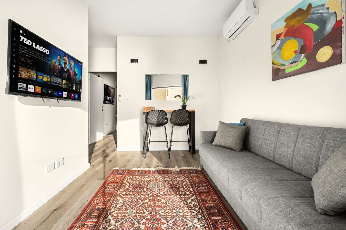 A modern living room with a grey sofa, a patterned rug, a TV mounted on the wall, and a painting.