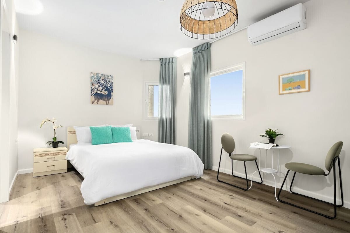 A modern bedroom with a large bed, a desk, two chairs, a small table, and a hanging light fixture.