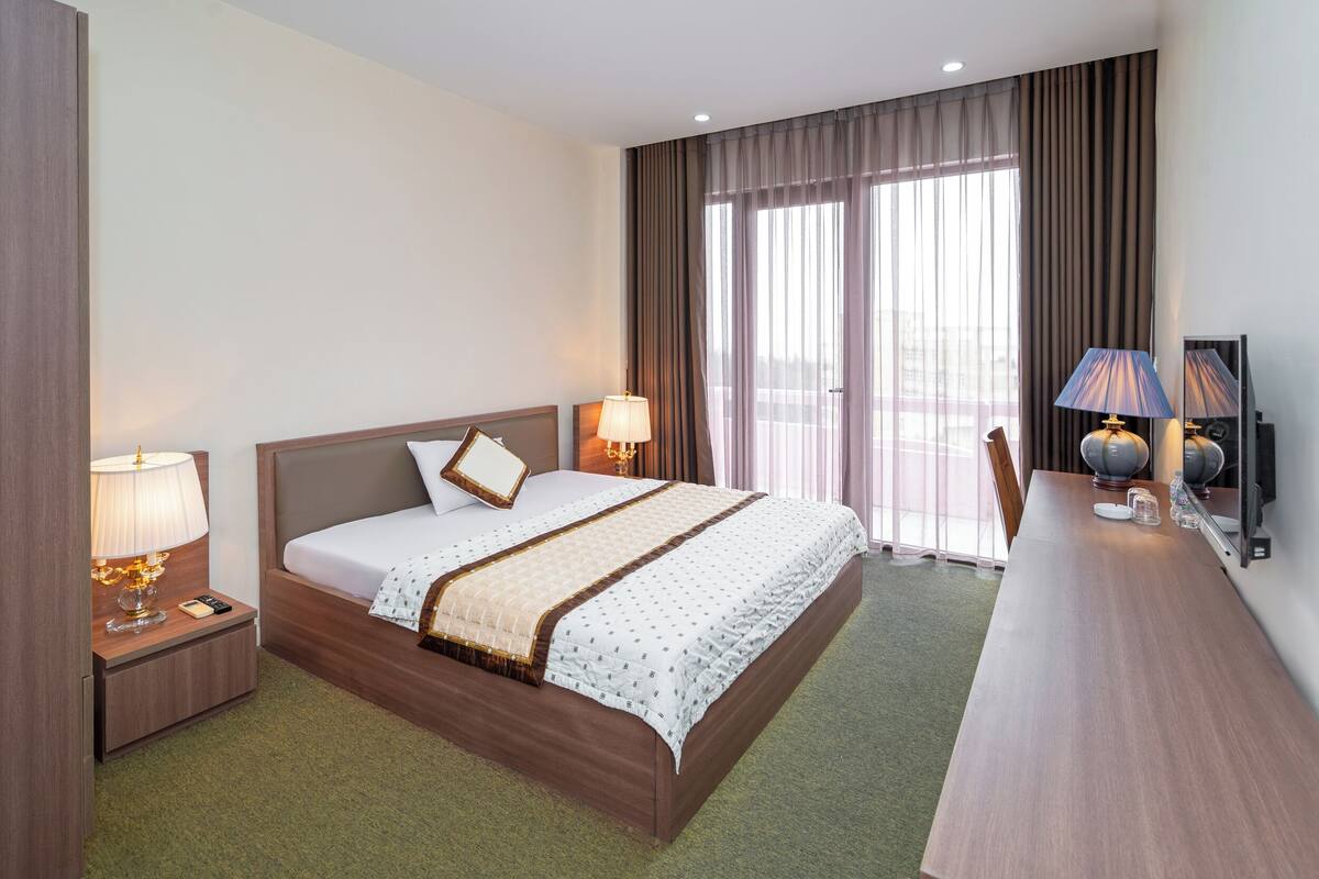 Une chambre d’hôtel avec un grand lit, deux lampes de chevet, un bureau avec une lampe et une vue sur l’extérieur grâce à de grandes fenêtres.