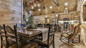 A rustic wooden interior with a dining table, chairs, a rocking chair, a Christmas tree, and a kitchen area. - The Liberty Lookout II | Ellijay, Ga (Ellijay)