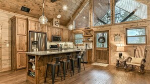 A rustic cabin interior with wooden walls, a large kitchen island, bar stools, and a rocking chair. - The Liberty Lookout II | Ellijay, Ga (Ellijay)
