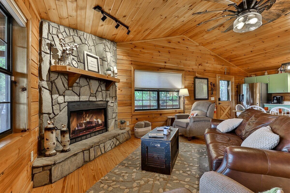 A cozy cabin interior with a stone fireplace, wooden walls, a leather sofa, and a dining area.