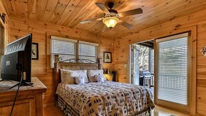 A wooden cabin bedroom with a bed, a ceiling fan, a TV on a wooden table, and a window with blinds.