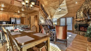 A spacious wooden cabin interior with a dining area, kitchen, fireplace, and a stone accent wall.