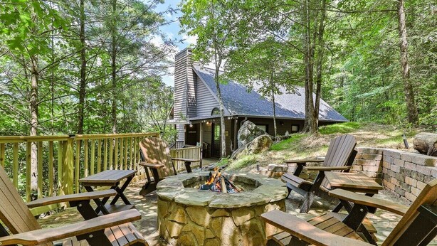 A stone fire pit surrounded by wooden Adirondack chairs, a wooden deck, and a cabin with a sloped roof.