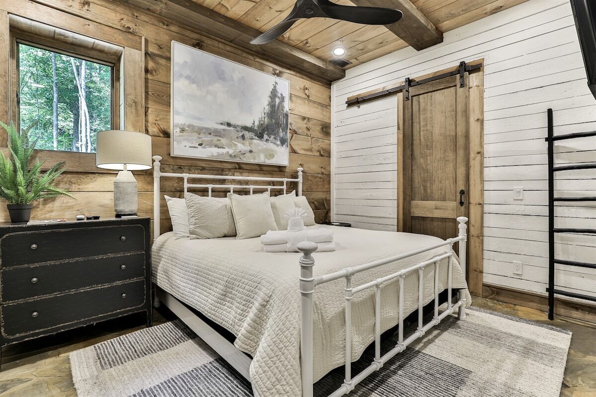 Een slaapkamer met een wit bed, een houten dressoir, een schilderij en een raam met uitzicht op bomen.