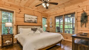 A wooden cabin bedroom with a bed, two bedside tables, a ceiling fan, and large windows. - Barn Owl`s Bliss | Cherry Log, GA (Ellijay)