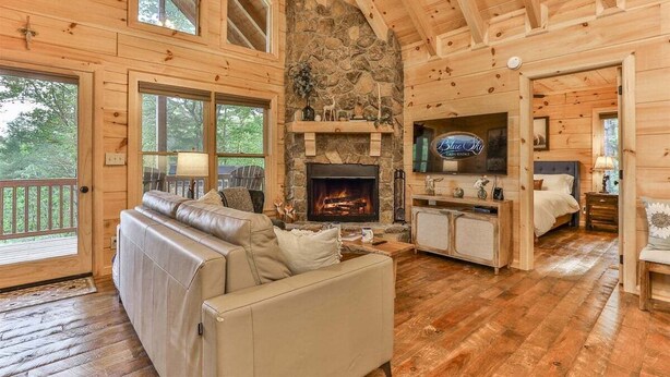 A log cabin interior with a stone fireplace, leather sofa, wooden floors, and a stone wall. - Barn Owl`s Bliss | Cherry Log, GA (Ellijay)