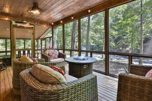 Un espace extérieur aménagé, abrité et doté de mobilier en osier, d’un foyer et de grandes fenêtres offrant une vue sur la forêt.