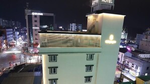 Un hôtel à plusieurs étages, avec un panneau lumineux sur le toit, un parking où sont garées plusieurs voitures, et une vue sur le paysage urbain environnant la nuit.