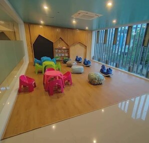 Relax in this spacious room with a high ceiling, featuring a large window offering a view of the outdoors and a cozy seating area with a bookshelf and play area.