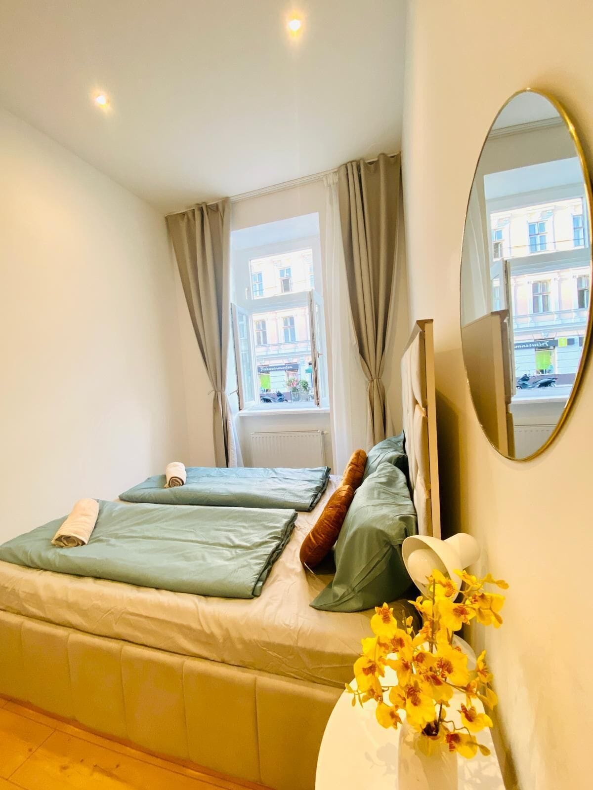 Un dormitorio con una cama, un espejo, una ventana con cortinas y un florero con flores.
