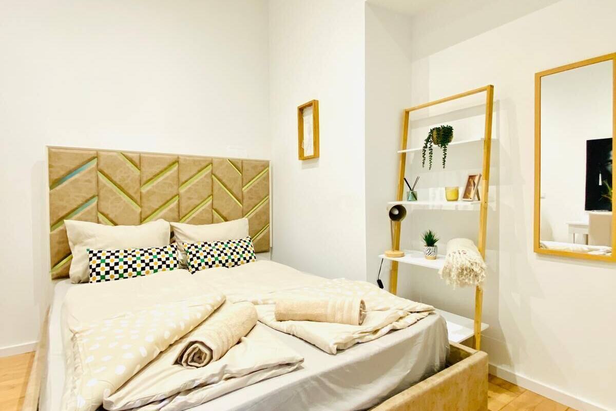 A modern bedroom with a bed, a bedside table, a mirror, and a small shelf with decorative items.
