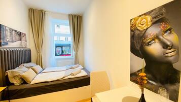 Una habitación de hotel moderna con cama, una mesa con un florero y copas de vino, un cuadro en la pared y una ventana con cortinas.