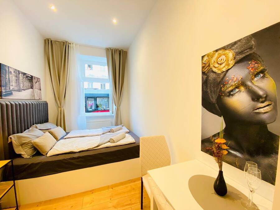 Una habitación de hotel moderna con cama, una mesa con un florero y copas de vino, un cuadro en la pared y una ventana con cortinas.