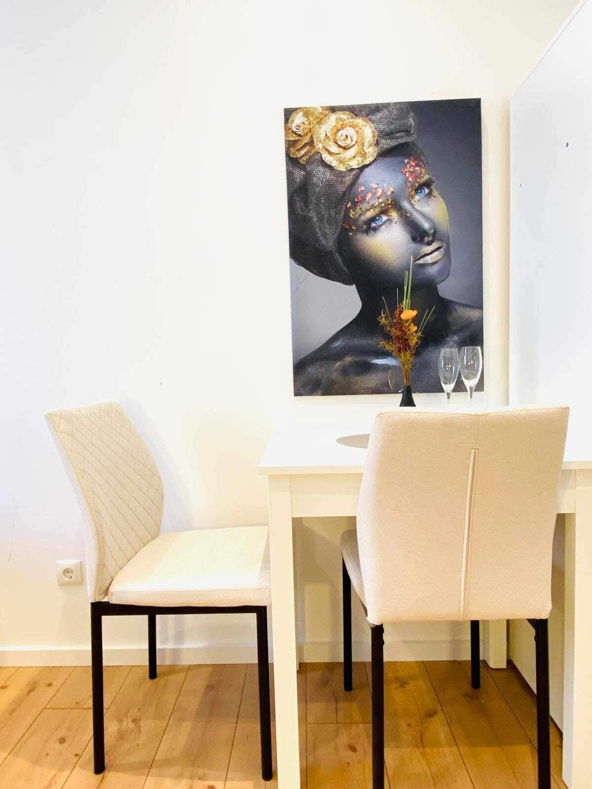Un comedor con una mesa blanca y dos sillas, un jarrón con flores y dos copas de vino.