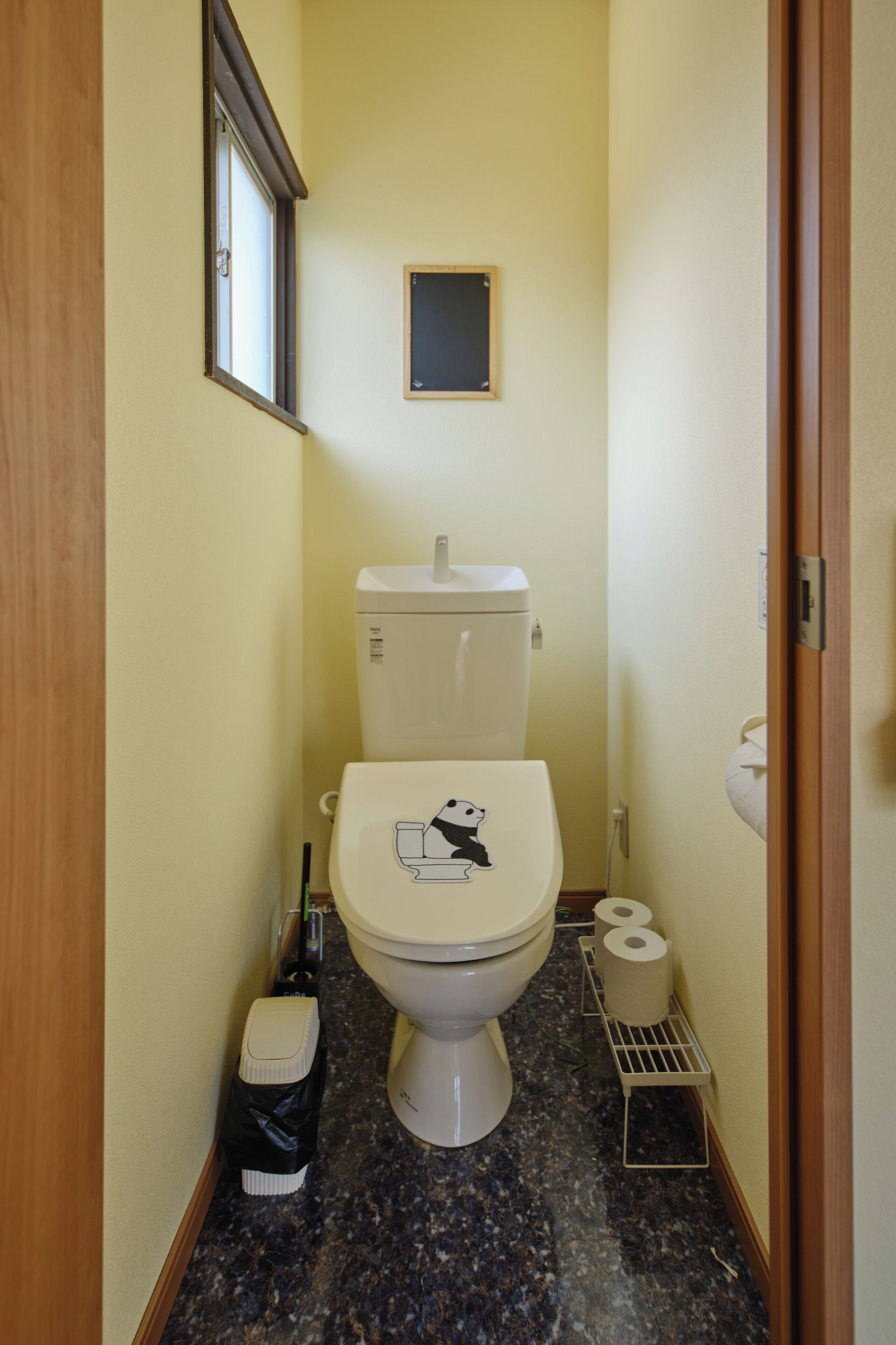 Casa de banho pequena com sanita, janela, porta-papel higiénico e um contentor de lixo.
