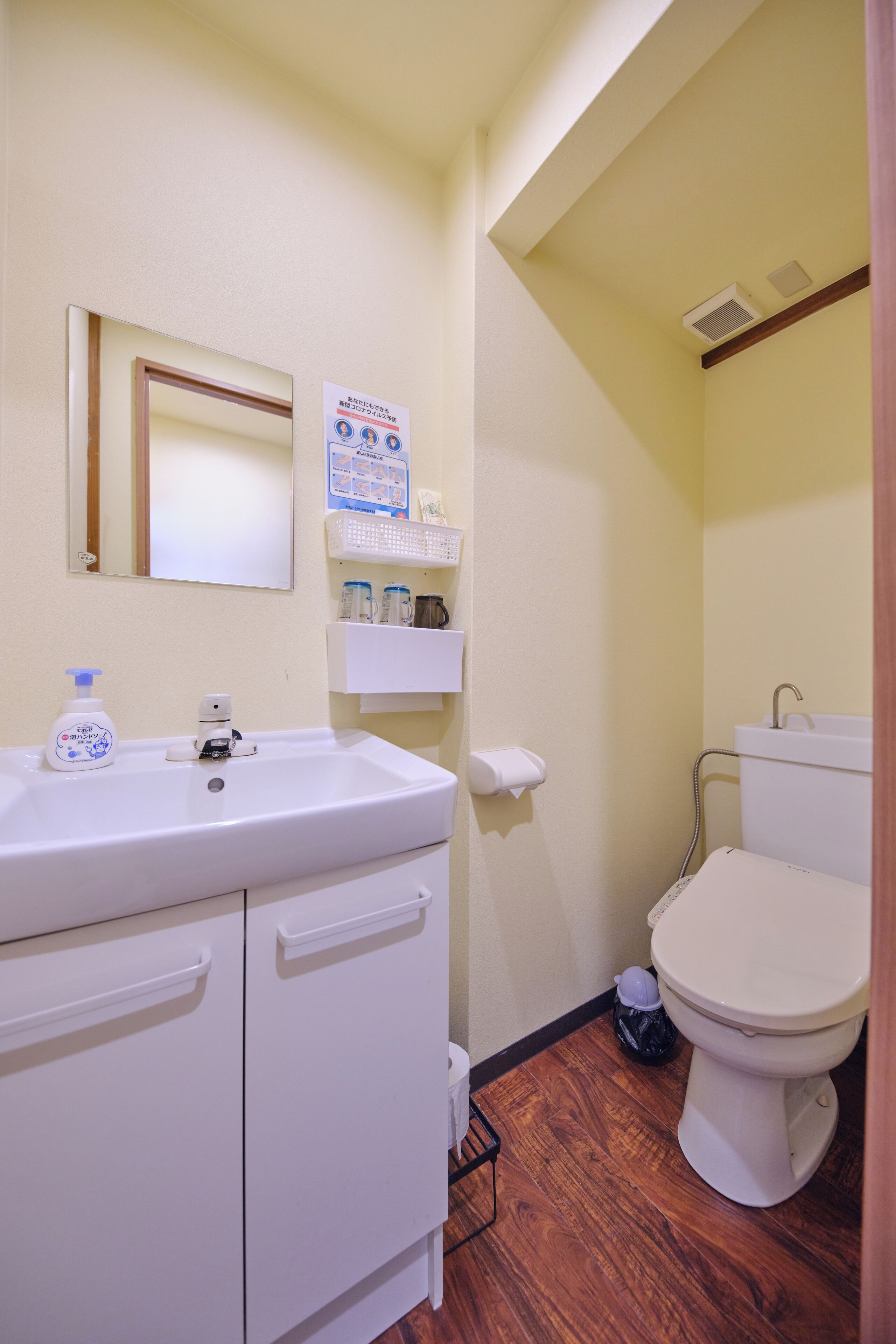 A bathroom with a white sink, mirror, and toilet. There are shelves with toiletries and a small trash bin.