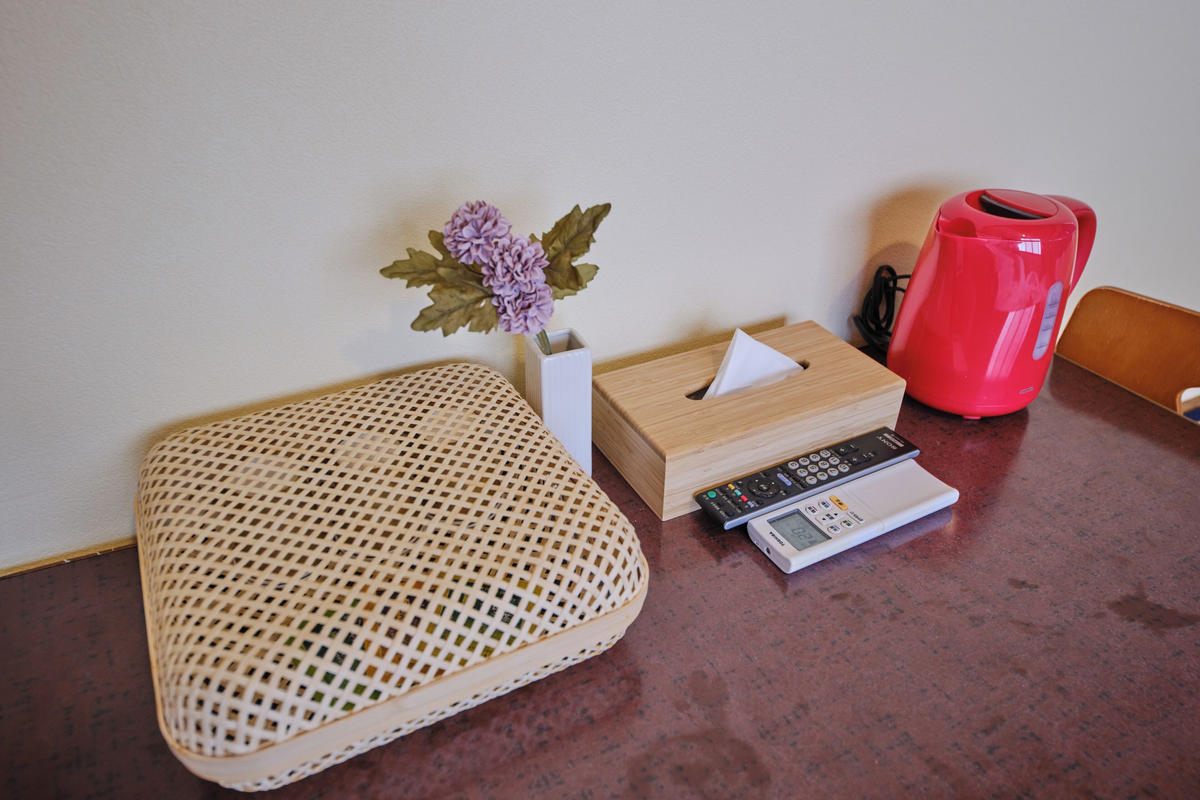 A wicker stool, a tissue box, a remote control, and a red kettle on a kitchen counter.