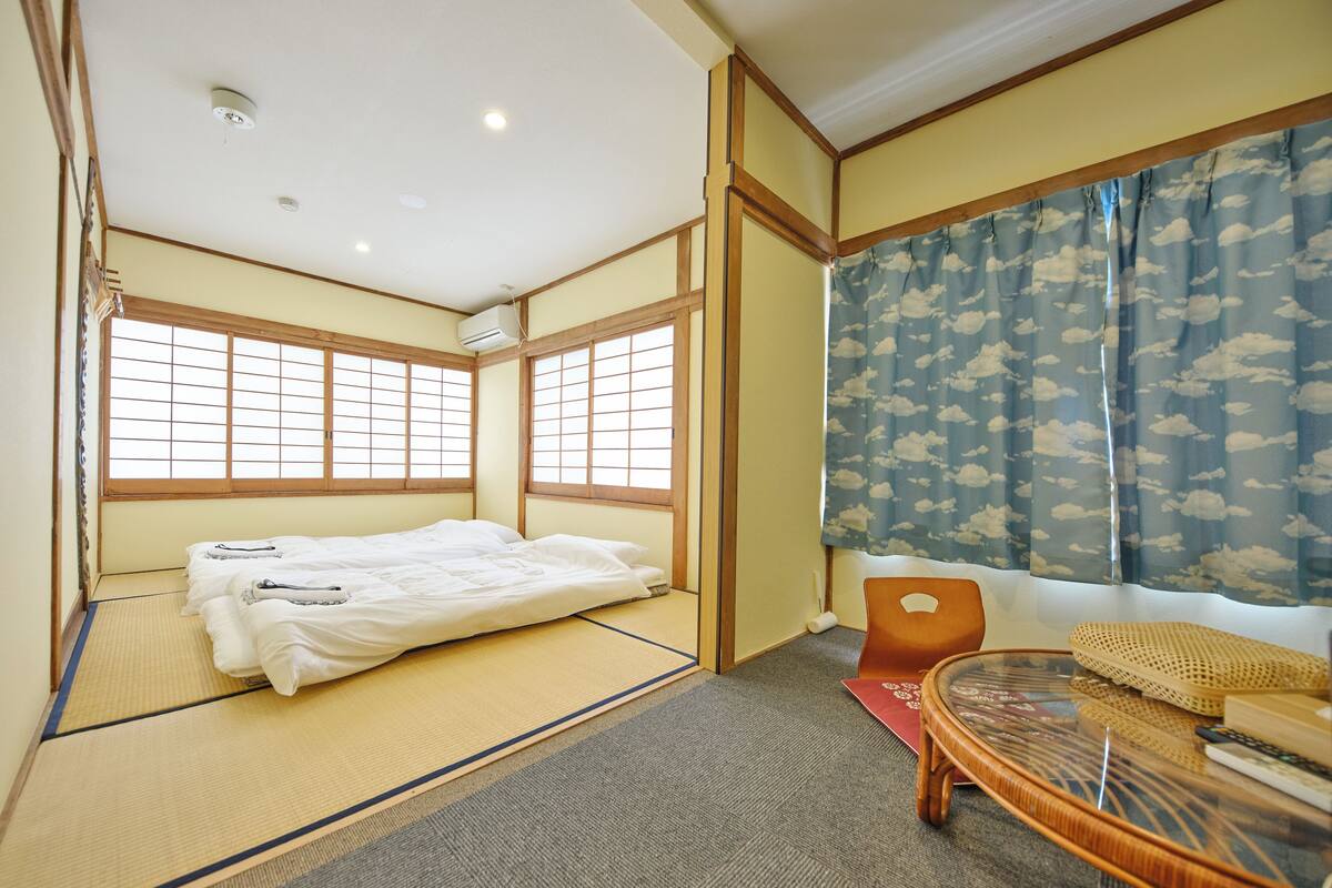 A traditional Japanese room with tatami flooring, a futon bed, a low table, and a sliding door with a cloud-patterned curtain.