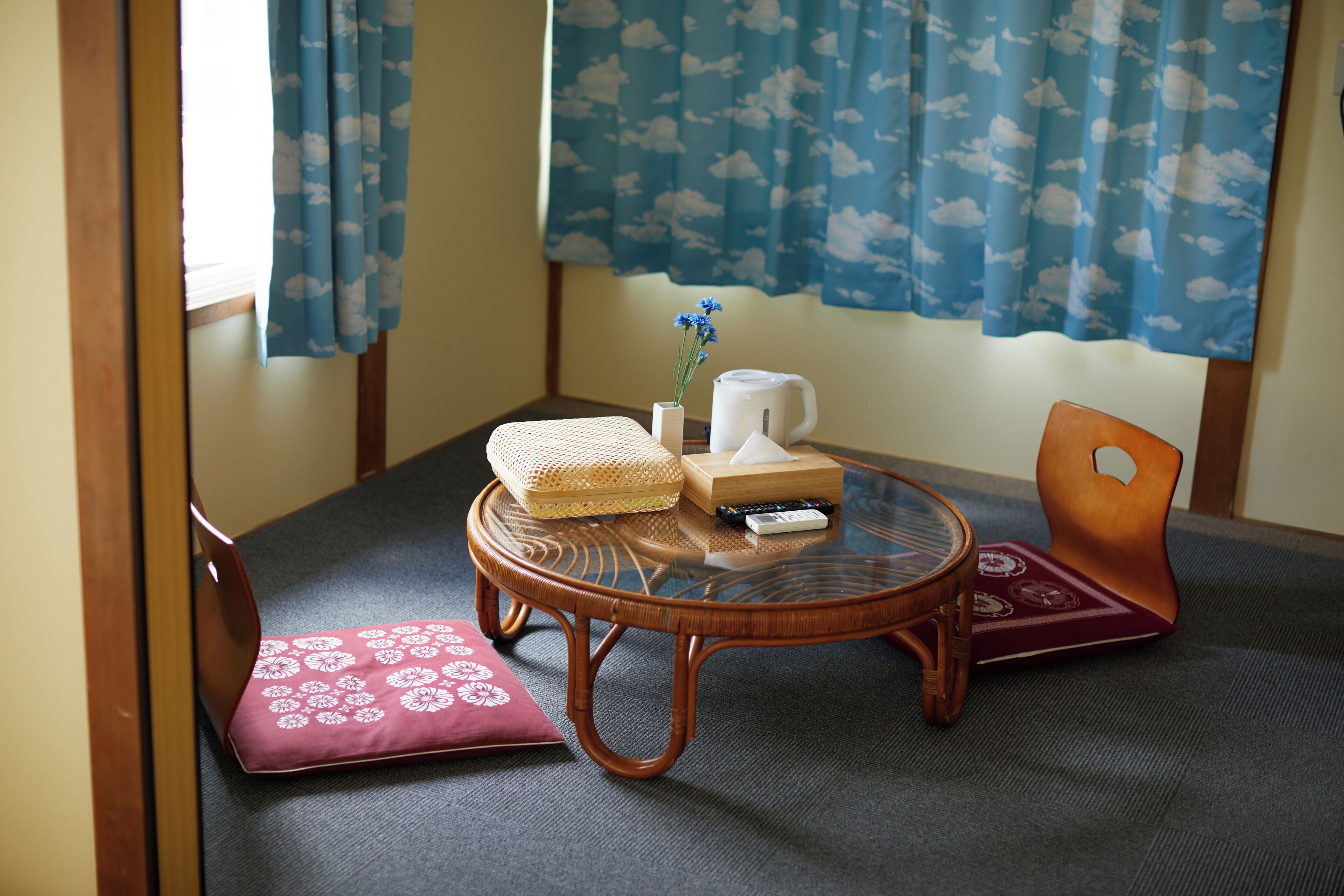 Una habitación con suelo de tatami, una mesa baja con una silla de mimbre, un cojín, un jarrón pequeño con flores, una tetera blanca y un control remoto.