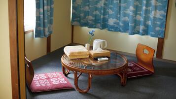 Una habitación con suelo de tatami, una mesa baja con una silla de mimbre, un cojín, un jarrón pequeño con flores, una tetera blanca y un control remoto.
