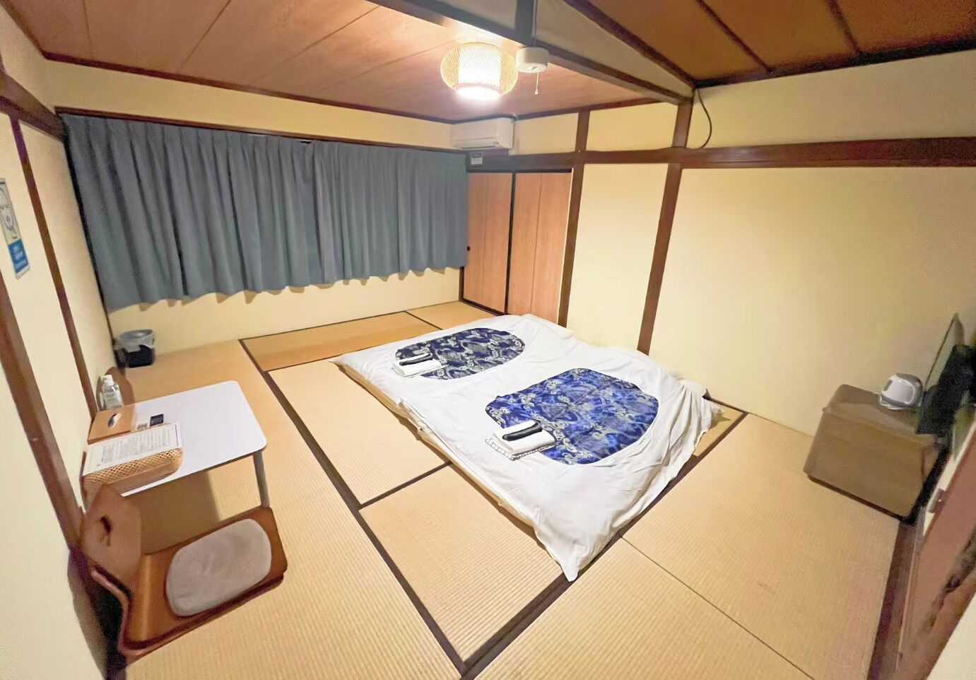 A room with tatami flooring, a bed with a blue and white bedspread, a small desk with a chair, and a window with a curtain.