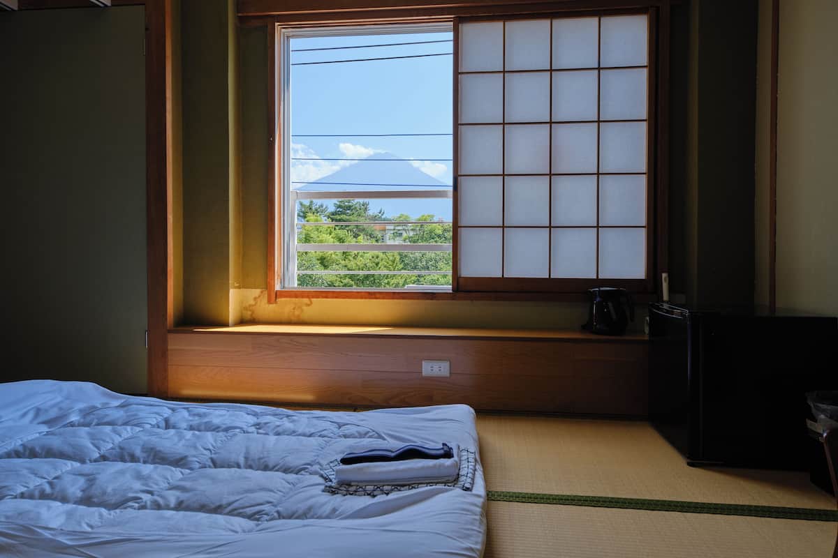 A room with a bed, a window with a view of greenery and a building, a small table with a kettle, and a black cabinet.