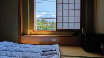 A room with a bed, a window with a view of greenery and a building, a small table with a kettle, and a black cabinet.