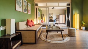 A modern living room with a sofa, a coffee table, and a television. There is a dining area with a table and chairs. The room has green walls and a large window. - Kssema (Mandrem)