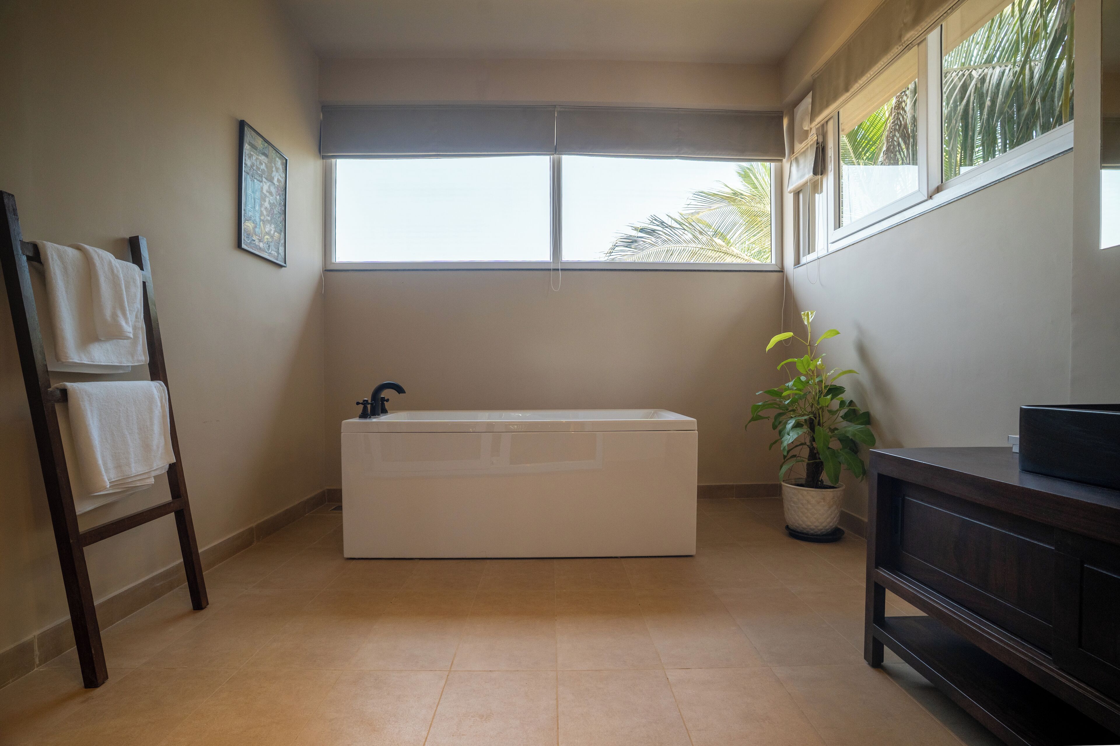 A bathroom with a bathtub, a towel rack, a sink, a potted plant, and a window with a view of palm trees.