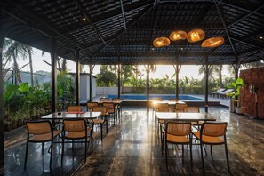 Un área de comedor al aire libre con mesas y sillas, una piscina al fondo y una estructura de techo cubierta.