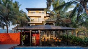 A two-story building with a pool and outdoor seating area, surrounded by palm trees. - Kssema (Mandrem)