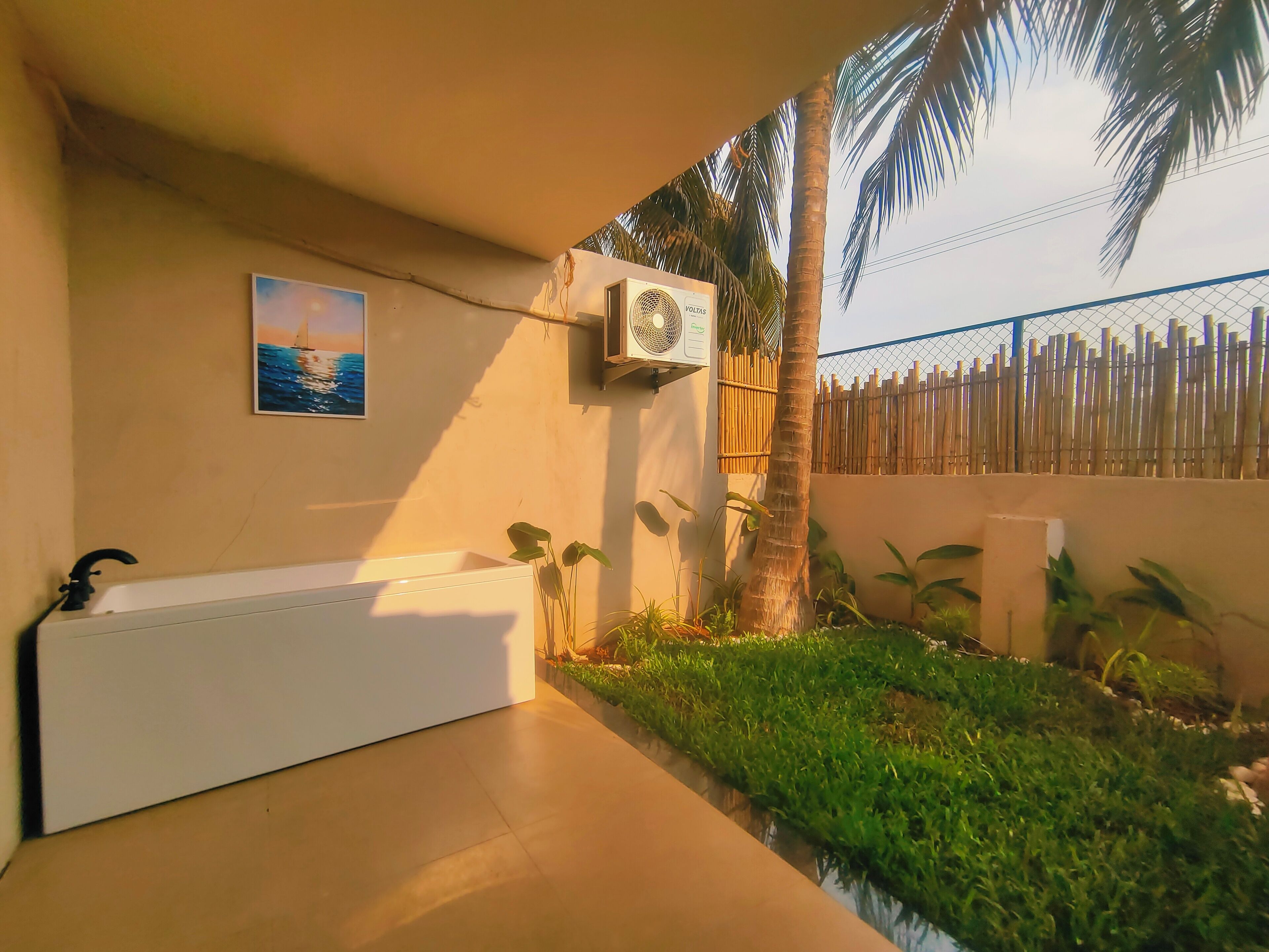 A bathroom with a bathtub, a wall-mounted air conditioner, a painting of a sunset over the ocean, and a bamboo fence.