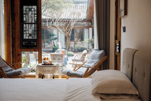 A room with a bed, two lounge chairs, a small table with flowers, and a view of a garden and building outside. - Jinfuyi hotel (Chengdu)