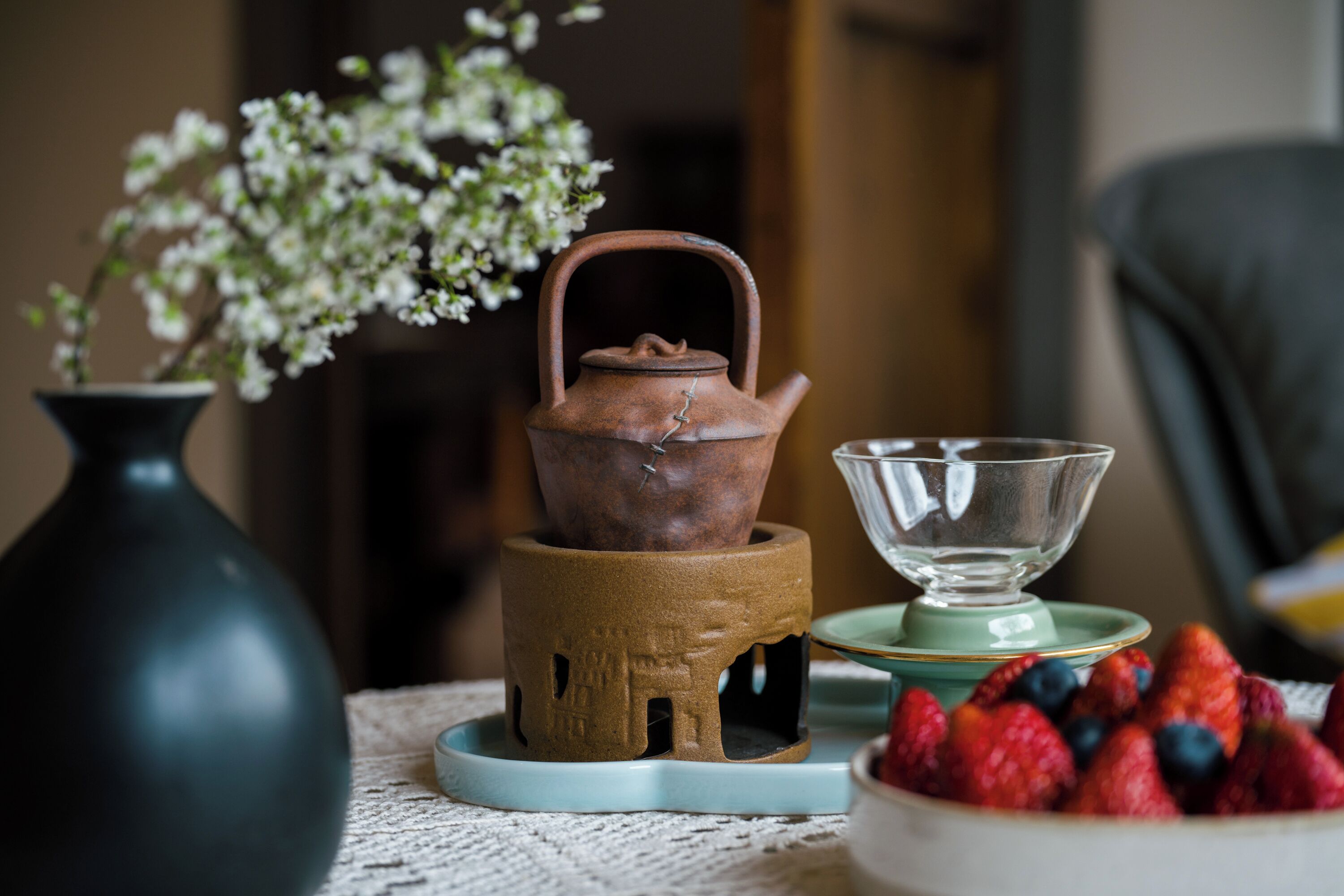 A tea set with a teapot and cup on a table, accompanied by a vase with flowers and a bowl of fruit.
