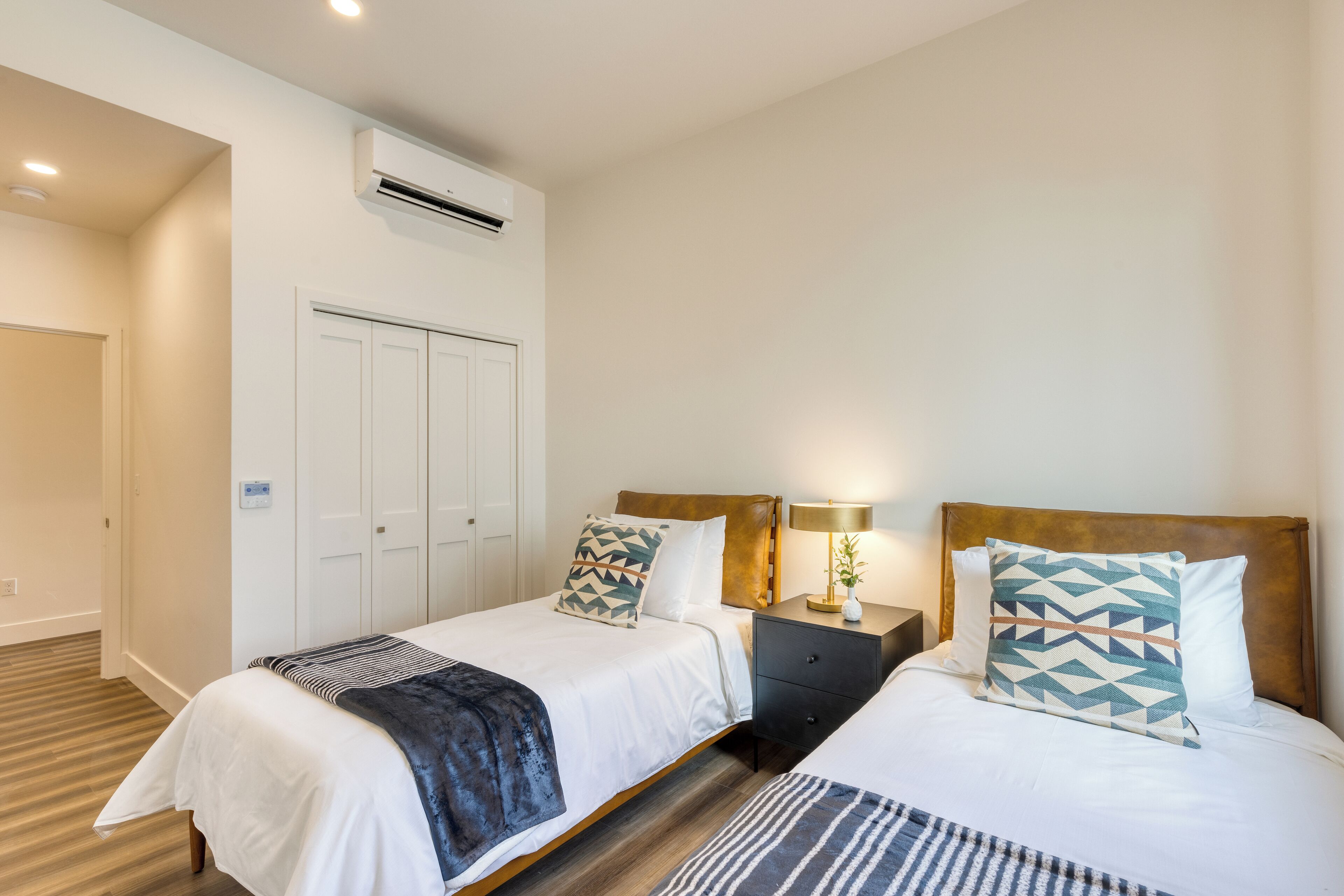 A hotel room with two beds, a wardrobe, an air conditioning unit, and a nightstand with a lamp.