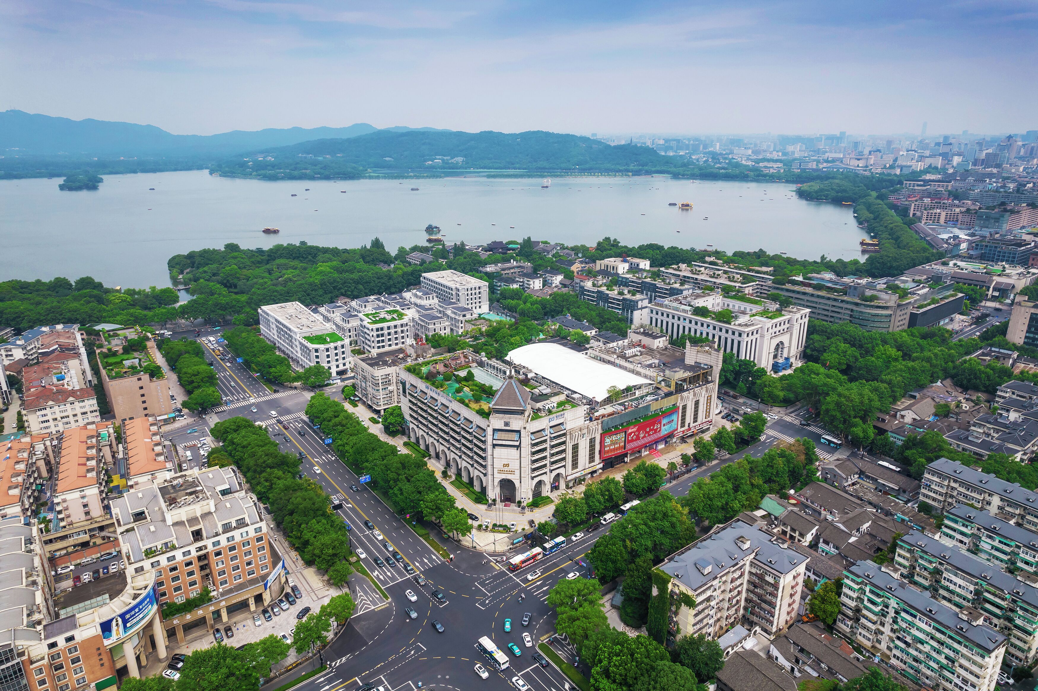 An aerial view of a cityscape with a prominent building featuring a green rooftop, surrounded by dense urban development and a large body of water in the background.
